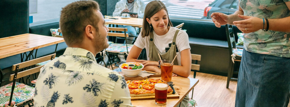 Banner of happy couple talking and ordering food to waiter in restaurant while are sitting at table with pizza, salad and drinks and another customer is sitting in the background.