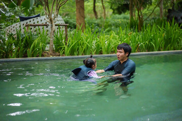 father teaching toddler girl swimming in pool and wearing life jacket