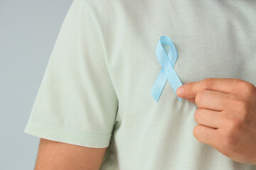 Young man with blue ribbon on light background, closeup. Prostate cancer awareness concept