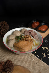 Hearty Indonesian Bakso soup with tofu and beef in a bowl, set on a dark table with rustic decor like pine cones and burlap.