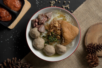 Flat lay of authentic Bakso, a traditional Indonesian soup with meatballs and tofu, on a dark rustic background with pine cones and spices.