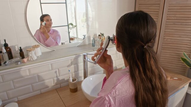 Young woman with dark long hair standing in front of round mirror in bathroom and using electric massager while making facial massage after morning hygiene