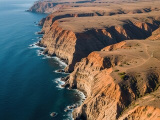 Aerial image of a coastal cliffside dividing deep blue ocean and rugged dry land, high contrast geological forms
