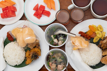 Close-up of traditional Indonesian meal with rice, soup, fried foods, and fruits on a beige background. Ideal for local cuisine or food culture content.
