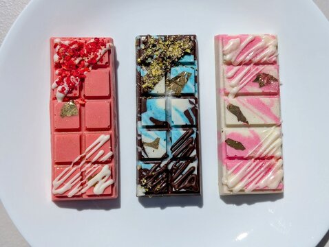 Assorted Flavors of Dubai Chocolate Bars with Decorative Toppings like Gold Flakes, Crushed Pistacchio, and White Cream on a White Plate
