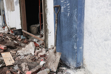 A shovel is standing and leaning against a blue wall, around it are the remains of a building that has been piled up in front of the door of a room that is being renovated
