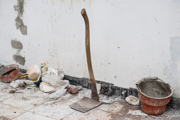 A hoe in front of a white wall with a dirty floor, around it there is a red bucket, a yellow ladle and a sack as well as bricks and some building debris, photographed from the right angle