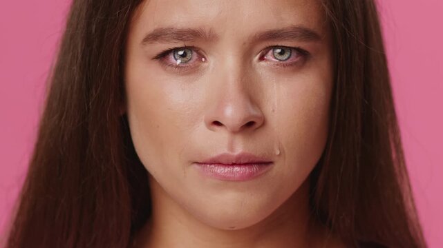A young Caucasian woman displays a deep emotional expression with tears in her eyes. The vibrant pink background emphasizes her feelings, creating a striking visual impact.