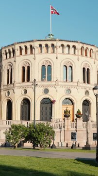 Vertical sunny exterior of Stortinget Parliament&rsquo;s circular chamber, Norway flag waving above, bright clear day, iconic Oslo architecture.