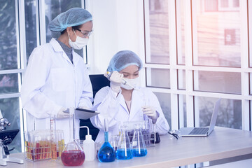 Two female scientists in lab coats and protective gear are performing an experiment, holding a flask with red liquid while observing and taking notes on a tablet.