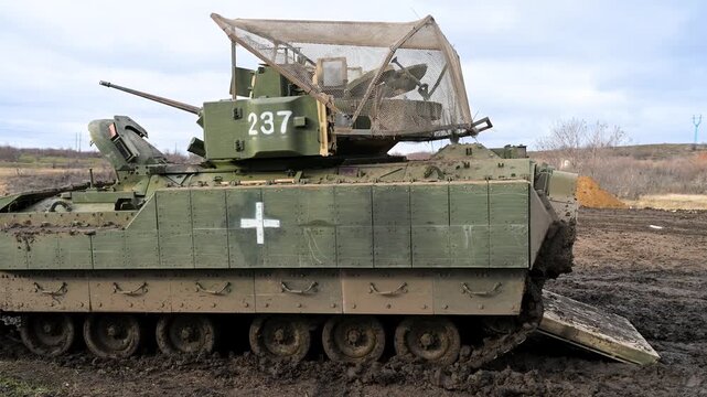 A US-made M2 Bradley, marked as a medical evacuation vehicle, navigates muddy terrain in Ukraine. It is fitted with a 'cope cage' for protection against drone attacks.