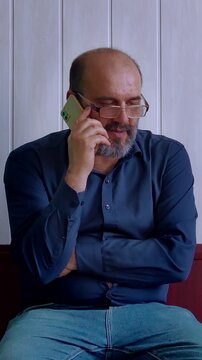 A serious and contemplative mood is captured as a bald man with a beard and glasses talks on a smartphone, looking down with a thoughtful expression. This shot is ideal for depicting business