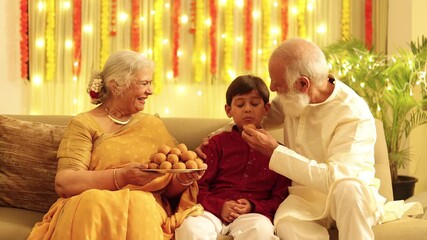 Happy indian grandparents and grandson enjoy eating laddoo or laddu sweet and celebrating diwali at home . Deepavali celebration with Lighting decoration. 4k - Powered by Adobe
