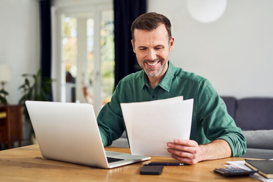 Smiling man working from home using laptop holding documents