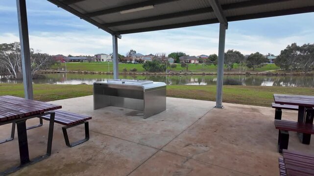 Covered picnic shelter with electric barbecue and timber tables at Navan Park, Melton, Melbourne Australia. Overlooking a peaceful lake and suburban home, scenic spot for outdoor gatherings.