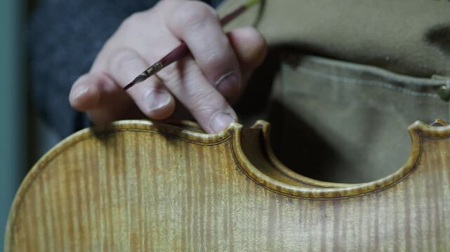 Close-up of a luthier hand-painting varnish onto a violin's ribs, capturing a precise moment in the antiquing process inside a workshop.