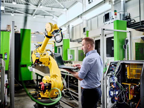 Technician using laptop at robotic arm in modern factory