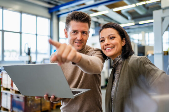 Smiling businessman holding laptop discussing about machine installation with businesswoman in industry