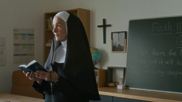 Medium shot of senior Caucasian nun in veil reading Bible while having religious lesson in modern classroom with crucifix on wall and holy quotes on chalkboard