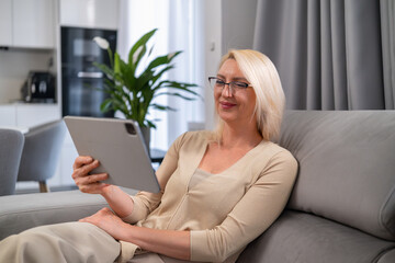 Calm mature woman sitting comfortably on sofa, reading the latest news on her tablet in a peaceful home environment. Digital lifestyle and information.