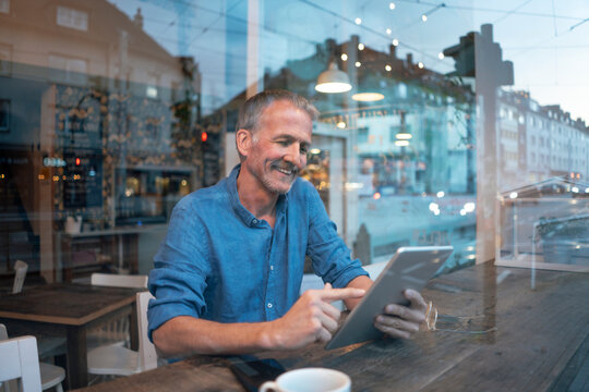 Smiling businessman sitting at table in front of window and using tablet PC at cafe