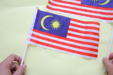 Close-up of two hands holding Malaysian flags on a light yellow background, symbolizing patriotism...