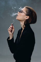Obraz premium Woman smoking electronic cigarette with stylish glasses and black jacket exhaling vapor in gray studio background