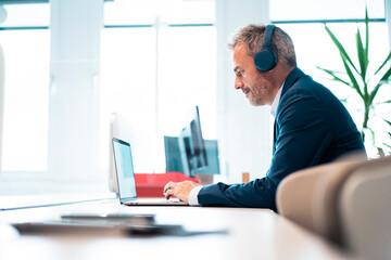 Businessman wearing wireless headphones using laptop at desk in workplace
