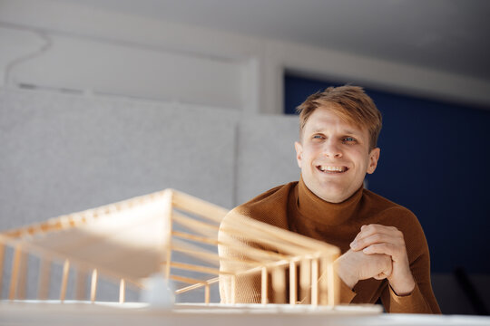 Smiling architect with hands clasped sitting by leaf shaped wooden model in office