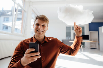 Happy businessman holding mobile phone pointing at levitating cloud network in office