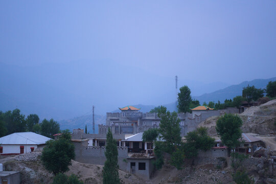 A remote village nestled in a mountainous landscape, featuring traditional architecture and scattered trees under a hazy sky.