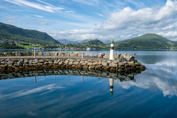 Obraz premium A peaceful harbor features a lighthouse and rocky shoreline under a blue sky, with mountains mirrored in the tranquil water.