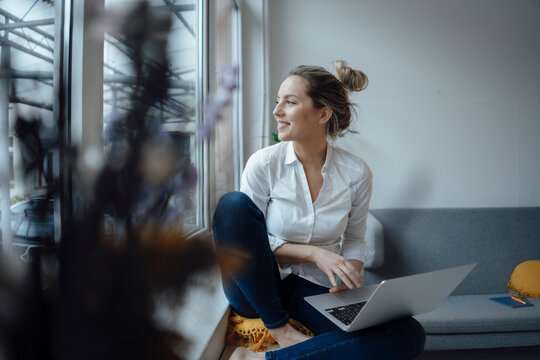 Smiling businesswoman with laptop sitting on widow sill in office