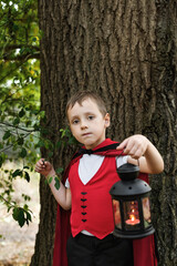 Child boy dressed as a vampire holding lantern near tree.