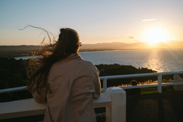 Woman watches Byron Bay sunset over ocean from lookout railing.