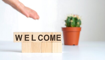 Wooden blocks spelling out "WELCOME" with a hand reaching above. A small cactus in a terracotta pot sits beside the blocks