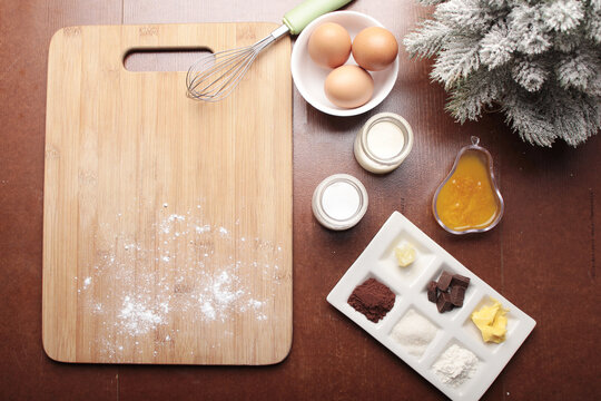 A top-down view shows baking ingredients including eggs, milk, flour, cocoa powder, chocolate, and butter laid out on a dark wooden surface next to a cutting board and a frosted pine branch - Powered by Adobe