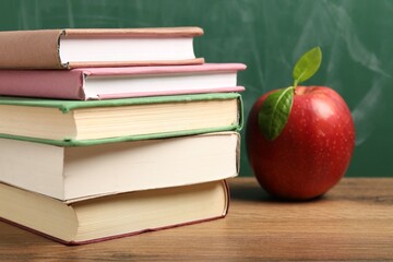 Different books and red apple on wooden table near blackboard, closeup