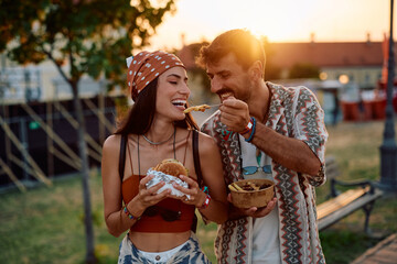 Happy couple having fun while sharing food at music festival at sunset.