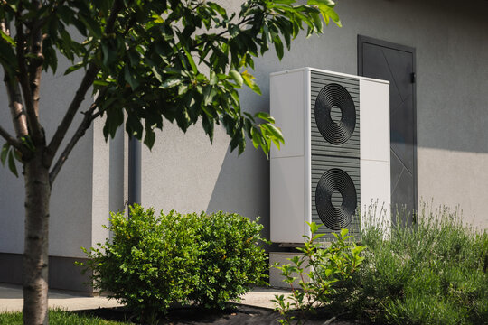 Modern outdoor heat pump unit installed near a residential house wall, surrounded by green shrubs and a tree in the landscaped yard