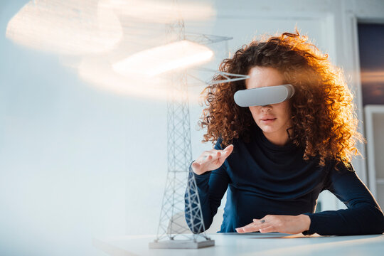 Businesswoman wearing virtual reality simulator gesturing by electricity pylon model on desk in office