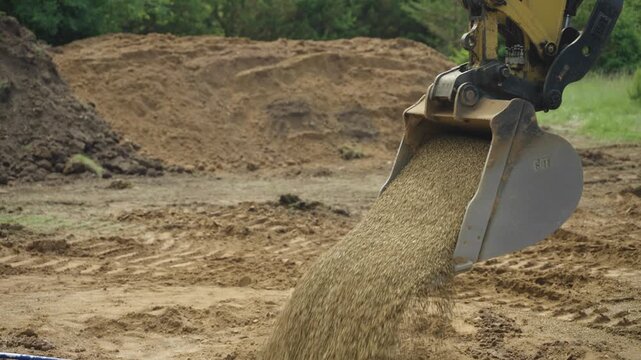Gravel pours from excavator bucket into trench for pool base layer prep.
