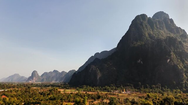Low drone pan in Vang Vieng, Laos showing towering karst cliffs, farmland, and scattered rural homes beneath rugged limestone peaks under soft morning light in dry season.