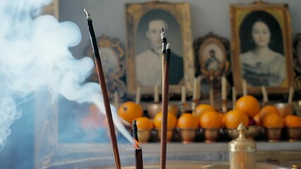 Incense sticks burn in a ceramic holder, emitting fragrant smoke that drifts upward over an ancestral altar adorned with framed portraits, oranges, and gently flickering candles