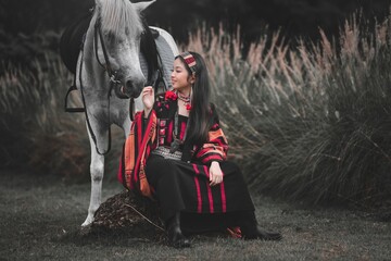 Young woman in traditional attire sitting beside gray horse outdoors