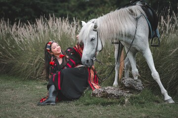 Young Woman in Traditional Attire Posing with White Horse Outdoors