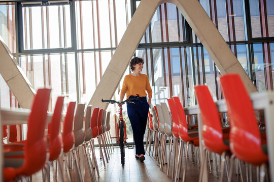 Smiling young businesswoman walking with bicycle near chairs in office cafeteria