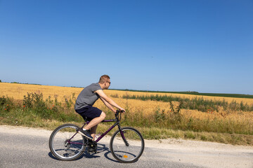 Fototapeta premium man cycling on a rural road beside a golden wheat field under summer sky