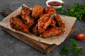 Baked chicken wings in a sweet chili sauce and sesame seeds on a wooden board on a dark concrete background.