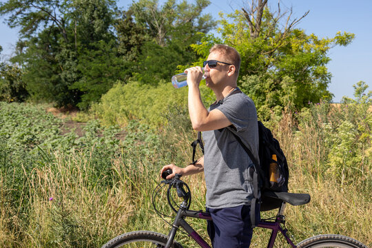 man drinking water from bottle while standing next to bicycle in nature